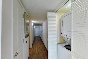 A laundry room with a washing machine and dryer, a wooden floor, and a hallway leading to other rooms. - Bay Watch Resort 1612 (North Myrtle Beach)
