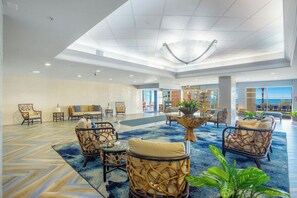 A spacious lobby with a high ceiling, a central seating area with wicker chairs and a glass-top table, and a view of the ocean through large windows. - Bay Watch Resort 1612 (North Myrtle Beach)