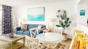 A living room with a sofa, a round glass table, and chairs. There is a plant, a clock, and a framed picture on the wall. - Bay Watch Resort 1612 (North Myrtle Beach)