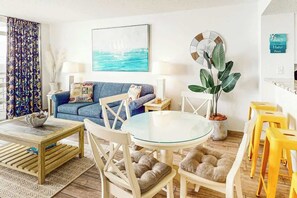 A living room with a sofa, a round glass table, and chairs. There is a plant, a clock, and a framed picture on the wall. - Bay Watch Resort 1612 (North Myrtle Beach)