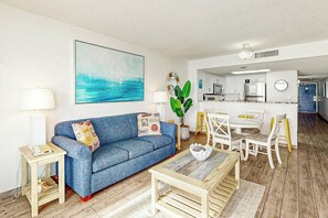 A living room with a blue sofa, a coffee table, and a dining area with white chairs. - Bay Watch Resort 1612 (North Myrtle Beach)