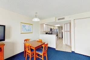 A room with a dining table and chairs, a television, a framed picture on the wall, and a kitchenette in the background. - Bay Watch Resort 1613 (North Myrtle Beach)