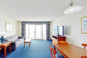 A hotel room with a sofa, a dining table, chairs, a TV, a lamp, and a view through the window. - Bay Watch Resort 1613 (North Myrtle Beach)