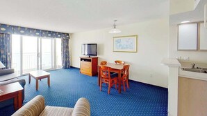 A hotel room with a sofa, a small table, a TV, a dining table with chairs, a kitchen area, and a large window with curtains. - Bay Watch Resort 1613 (North Myrtle Beach)