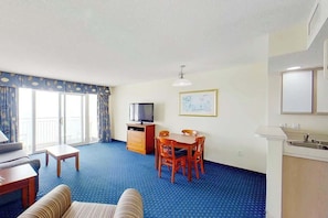 A hotel room with a sofa, a small table, a TV, a dining table with chairs, a kitchen area, and a large window with curtains. - Bay Watch Resort 1613 (North Myrtle Beach)