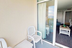 A balcony with a white chair and table, a glass door leading to a room with a bed and a small table. - Bay Watch Resort 1615 (North Myrtle Beach)