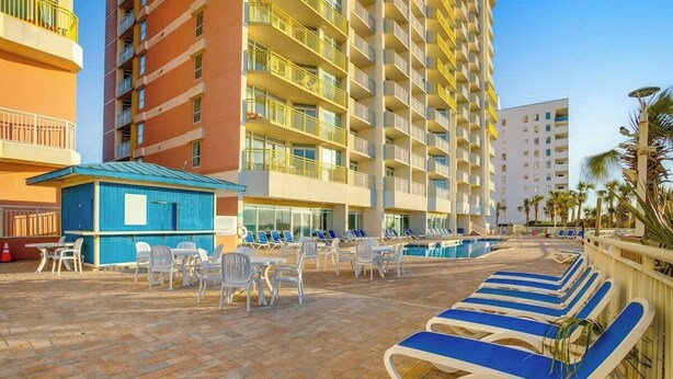 Ein Hotelpoolbereich mit Liegestühlen und Tischen, ein kleines blaues Gebäude sowie ein hohes Hotelgebäude mit Balkonen.