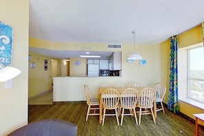 A dining area with a table and chairs, a kitchen in the background, and a window with curtains. - Bay Watch Resort 1702 (North Myrtle Beach)