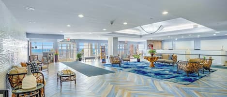 A spacious lobby with a herringbone floor pattern, seating areas with wicker furniture, and a view of the sea through large windows.