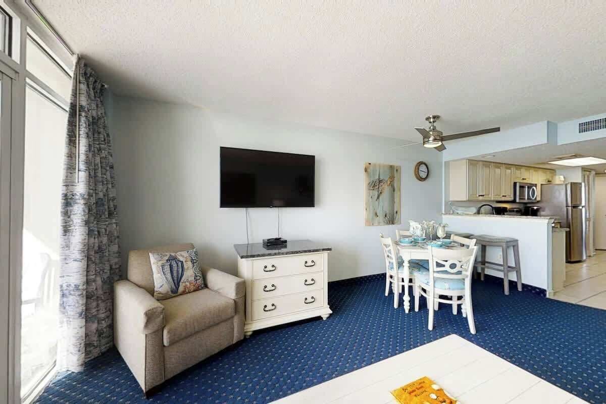 Habitación de hotel con sofá, televisor de pantalla plana, mesa de comedor con sillas, zona de cocina y un ventanal con cortinas.