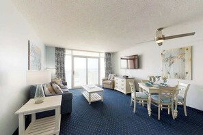 Une chambre d’hôtel avec un canapé, une table à manger avec des chaises, une petite table d’appoint, un ventilateur de plafond et une grande fenêtre.