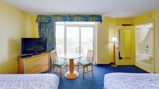 Une chambre d’hôtel équipée d’une télévision, d’une table à manger avec des chaises, d’un balcon avec vue, d’une salle de bain avec douche et d’un lit.