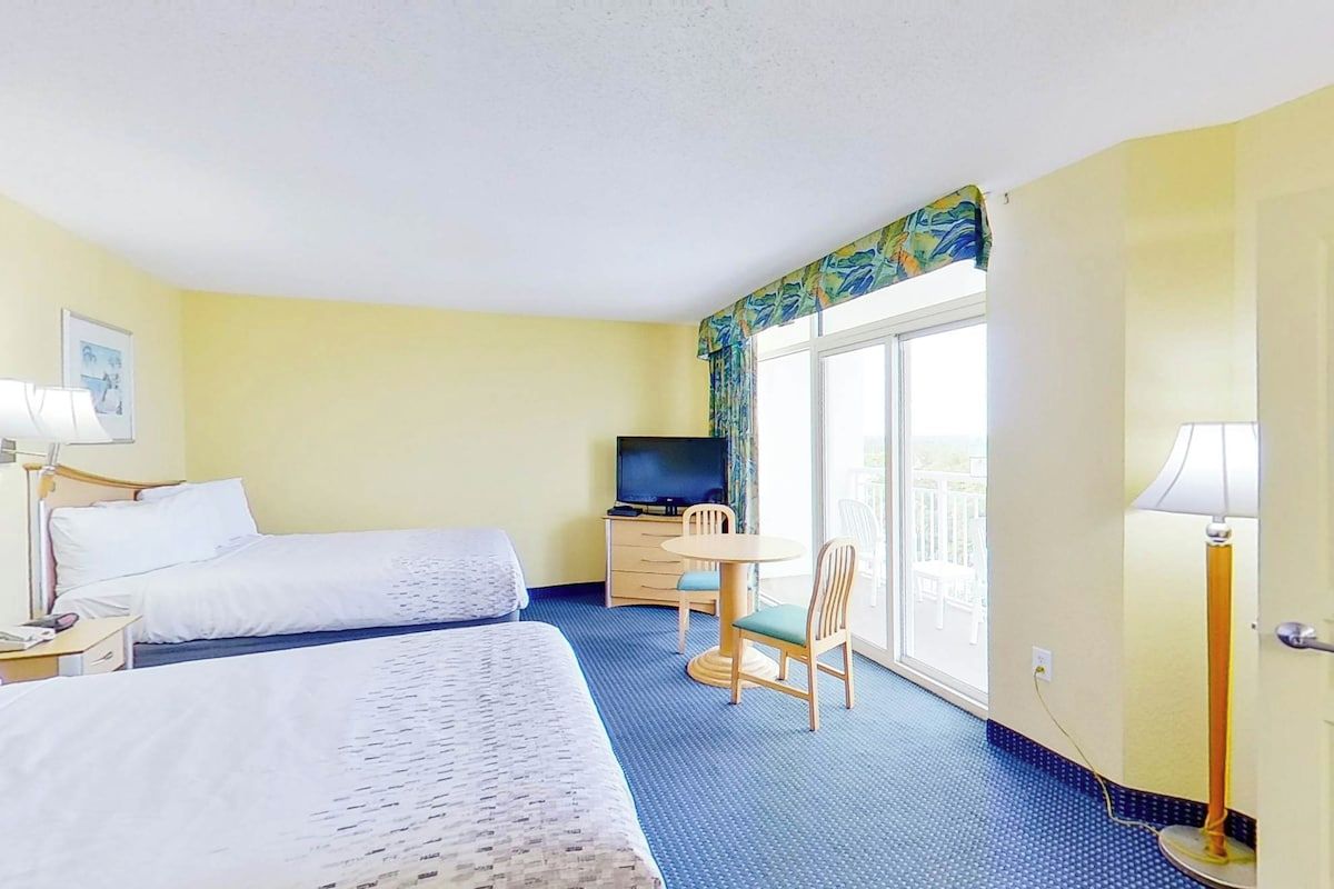Quarto de hotel com duas camas, televisão, escrivaninha e cadeira, além de janela com vista.