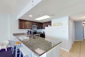 A kitchen with granite countertops, dark wooden cabinets, a stainless steel refrigerator, and a microwave. There are two bar stools and a white chair. - Bay Watch Resort 1805 (North Myrtle Beach)