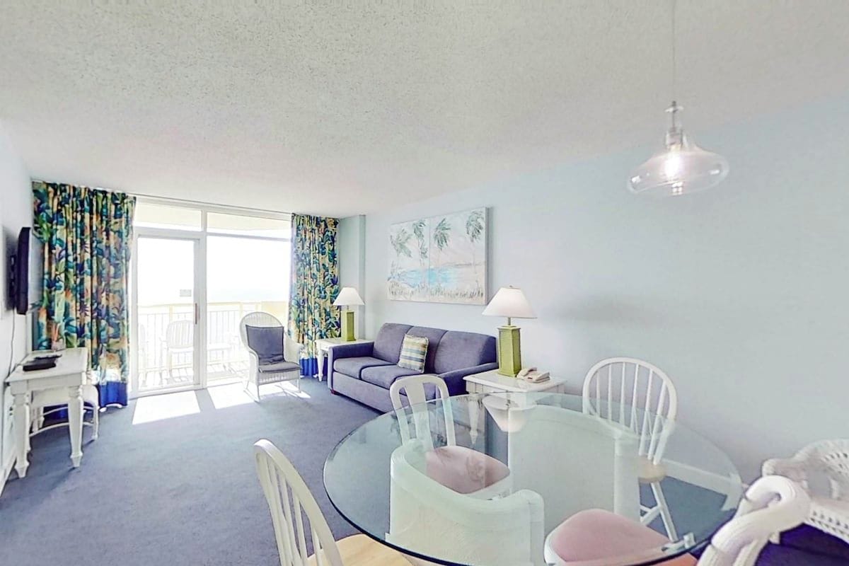 A living room with a glass table, chairs, a sofa, a television, and a balcony with curtains.