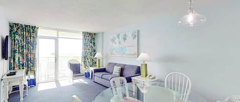 Olohuoneessa on lasipöytä, tuoleja, sohva, televisio ja parveke, jossa on verhot.