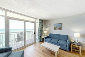 A room with a large sliding glass door leading to a balcony, a blue sofa, a white coffee table, and a small wooden side table with a lamp. - Bay Watch Resort 1820 (North Myrtle Beach)