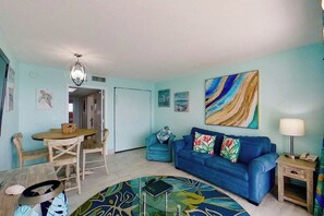 A living room with a blue sofa, a dining table with chairs, a patterned rug, and a large abstract painting on the wall. - Bay Watch Resort 1906 (North Myrtle Beach)