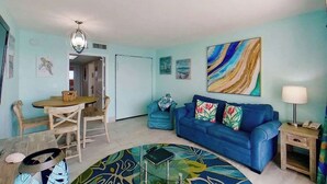 A living room with a blue sofa, a dining table with chairs, a patterned rug, and artwork on the walls. - Bay Watch Resort 1906 (North Myrtle Beach)