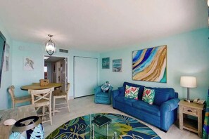 A living room with a blue sofa, a dining table with chairs, a patterned rug, and artwork on the walls. - Bay Watch Resort 1906 (North Myrtle Beach)