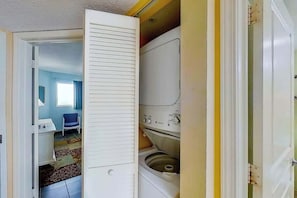 A laundry room with a washing machine and dryer, a chair, and a small table. - Bay Watch Resort 1919 (North Myrtle Beach)