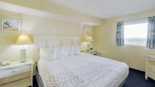 A hotel room with a large bed, two bedside tables with lamps, a window with curtains, and artwork on the walls. - Bay Watch Resort 1920 (North Myrtle Beach)
