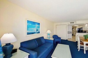 A living room with a blue sofa, a coffee table, a lamp, and a painting of a sailboat on the wall. - Bay Watch Resort 1920 (North Myrtle Beach)