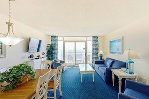 A room with a dining table, chairs, a sofa, a TV, a balcony with curtains, and a plant. - Bay Watch Resort 1920 (North Myrtle Beach)