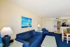 Habitació d'hotel amb un sofà blau, una taula petita, una làmpada, un quadre d'un veler i una cuineta al fons.