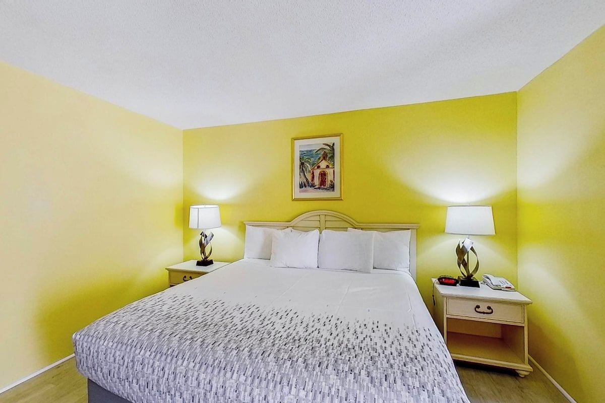 Habitación de hotel con cama, dos mesitas de noche con lámparas, una mesita auxiliar y un cuadro en la pared.