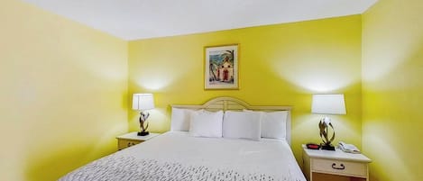 Habitación de hotel con cama, dos mesitas de noche con lámparas, una mesita auxiliar y un cuadro en la pared.