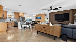 Eine moderne Küche mit einer zentralen Kochinsel, ein Essbereich mit einer Couch und Stühlen sowie ein Wohnzimmer mit Fernseher und Deckenventilator.