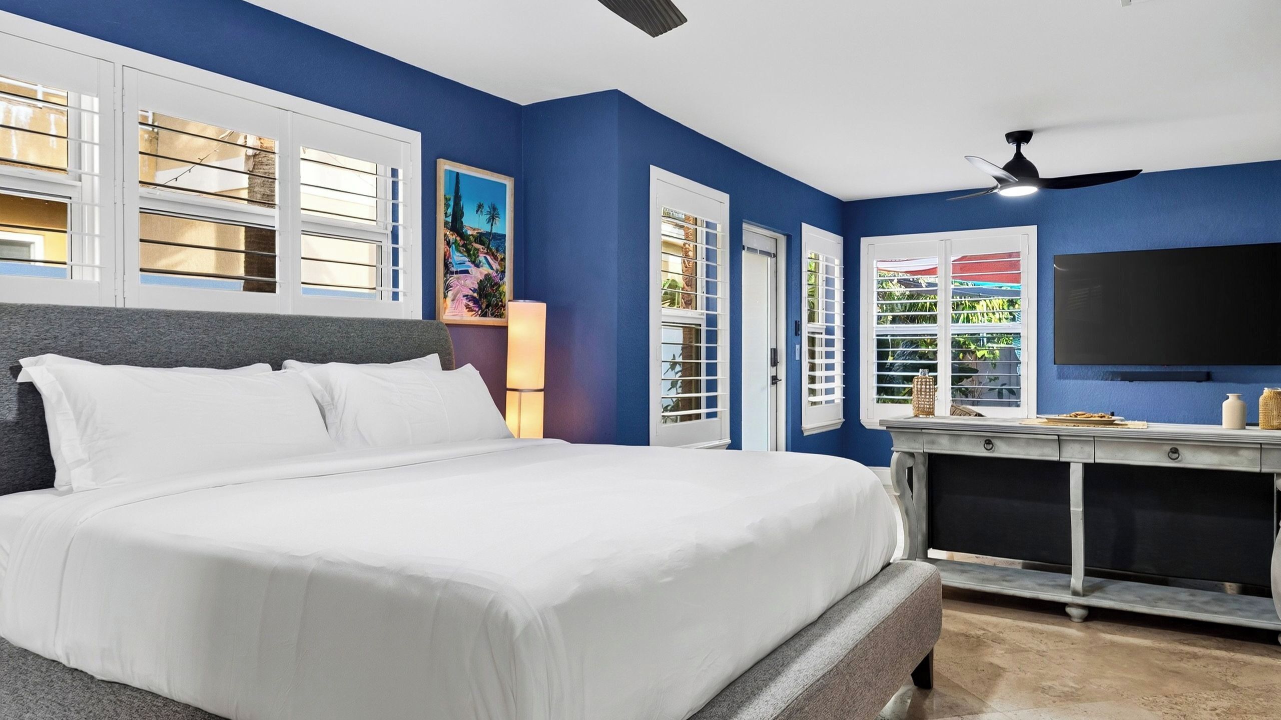 A bedroom with a large bed, a desk, a TV, and a ceiling fan.