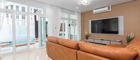 Una sala de estar moderna con un sofá de cuero, un televisor de pantalla plana montado en una pared de madera, y amplios ventanales correderos que dan a un espacio exterior.