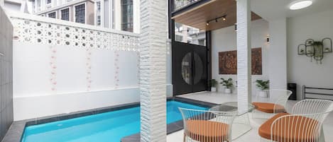 Un espace piscine intérieur moderne, avec une piscine à l’eau d’un bleu limpide, des murs blancs et un balcon offrant une vue sur les bâtiments environnants.