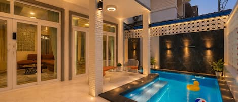 Un área de piscina moderna al aire libre con cerramiento de vidrio, zona de estar con sofá y sillones, y un muro decorativo con diseño geométrico.