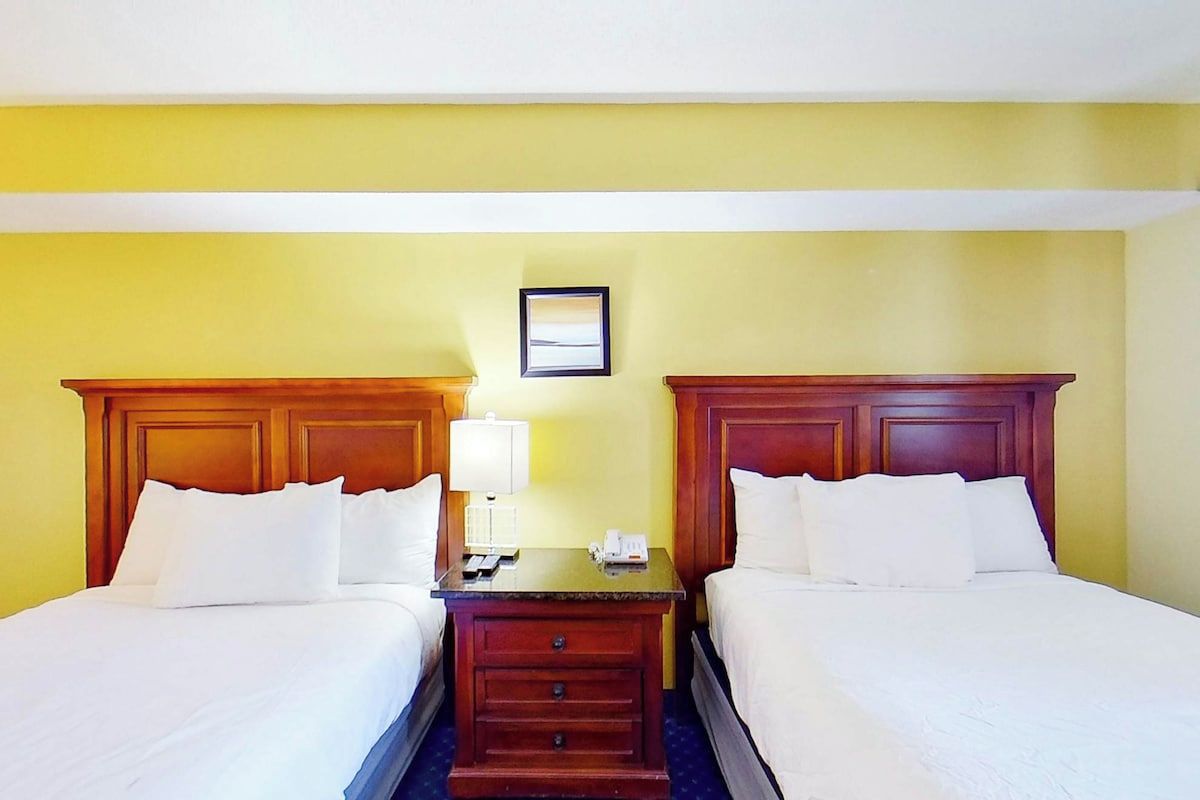 A hotel room with two beds, a nightstand, a lamp, and a framed picture on the wall.