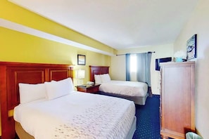 A hotel room with two beds, a wooden wardrobe, a desk with a lamp, and a window with curtains. - Bay Watch Resort 1936 (North Myrtle Beach)