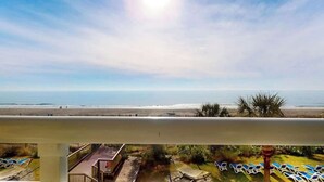 A balcony with a view of a beach, sun, and lounge chairs. - Camelot by the Sea 0307 (Myrtle Beach)