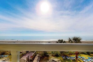 A balcony with a view of a beach, sun, and lounge chairs. - Camelot by the Sea 0307 (Myrtle Beach)