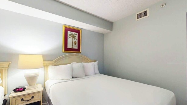 A hotel room with a bed, bedside tables, a nightstand with a lamp, and a wall-mounted picture. - Camelot by the Sea 0307 (Myrtle Beach)