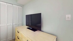 A room with a television mounted on a wall, a wooden dresser, and a set of white louvered closets. - Camelot by the Sea 0307 (Myrtle Beach)