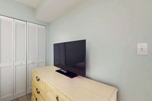 A room with a television mounted on a wall, a wooden dresser, and a set of white louvered closets. - Camelot by the Sea 0307 (Myrtle Beach)