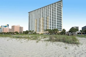 Un hôtel de grande hauteur au design moderne, situé près d’une plage de sable fin bordée de palmiers.