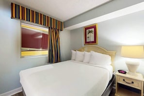 A hotel room with a large bed, a nightstand with a lamp, a framed picture on the wall, and a window with curtains. - Camelot by the Sea 0307 (Myrtle Beach)