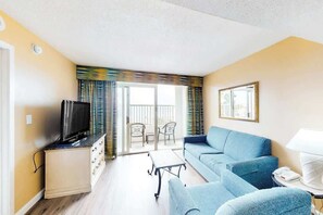 Ein Wohnzimmer mit einer Couch, einem Fernseher auf einem Schrank, einem Couchtisch und einem Balkon mit Stühlen.