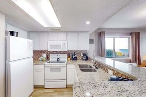 A modern kitchen with white cabinetry, a granite countertop, a built-in oven, and a window with curtains. - Camelot by the Sea 0317 (Myrtle Beach)