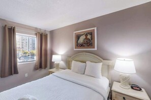 A hotel room with a bed, two bedside tables with lamps, a window with curtains, and a painting on the wall. - Camelot by the Sea 0317 (Myrtle Beach)