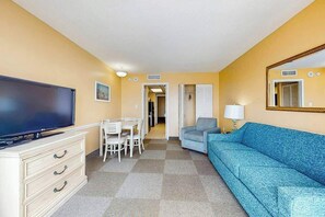 A hotel room with a television, a dining table with chairs, a sofa, and a mirror. - Camelot by the Sea 0916 (Myrtle Beach)