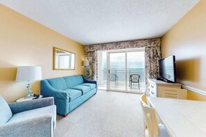 A hotel room with a sofa, a TV, a dining table, and a balcony with a view. - Camelot by the Sea 0916 (Myrtle Beach)